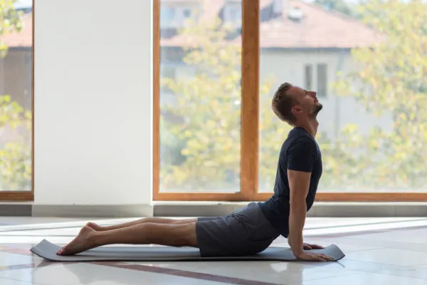 Man practicing Bhujangasana, or cobra pose, on a yoga mat in a bright studio with large windows and natural light.