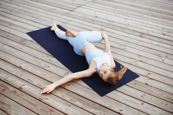 Woman doing a reclining supine twist pose on a navy yoga mat over a light wooden deck, stretching her torso.