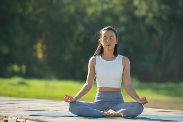Woman meditating in cross-legged lotus posture outdoors on a wooden deck, practicing Yoga and Longevity routine.