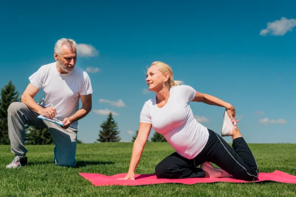 Senior woman guided by instructor while performing a leg stretch outdoors to support Yoga and Longevity goals.