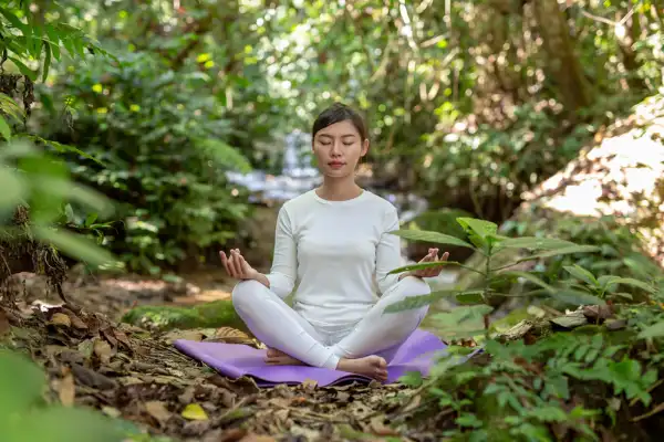 Woman meditating in lotus pose outdoors in a lush forest retreat, embracing holistic wellness through ayurveda and yoga.